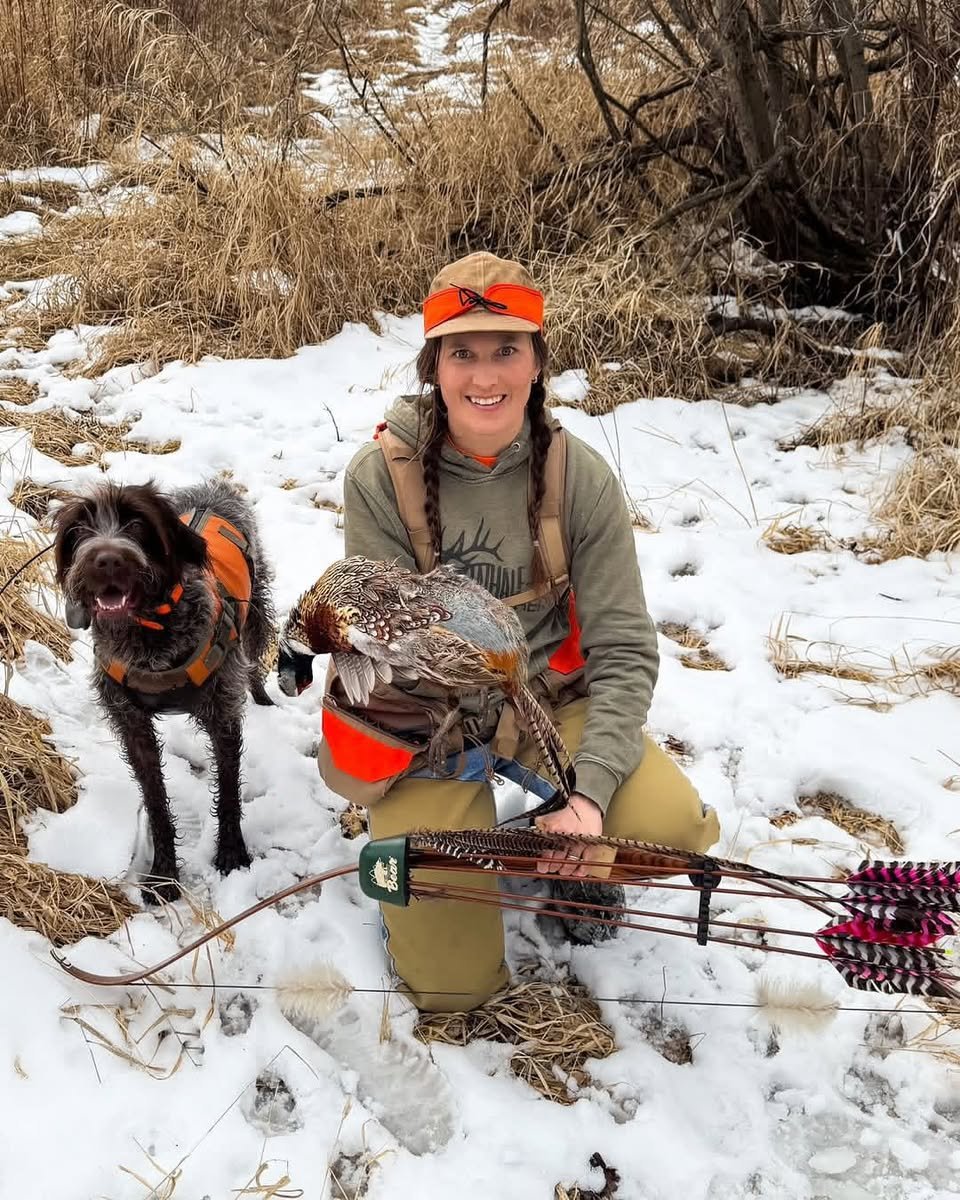 Female hunter in the snow with a pheasant, recurve bow, wood-grain feather-fletched arrows and her hunting dog